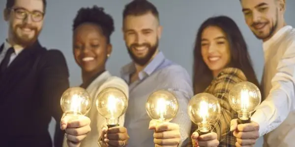 Diverse group of young businesspeople smiling and holding shining light bulbs to represent sharing a useful idea.
