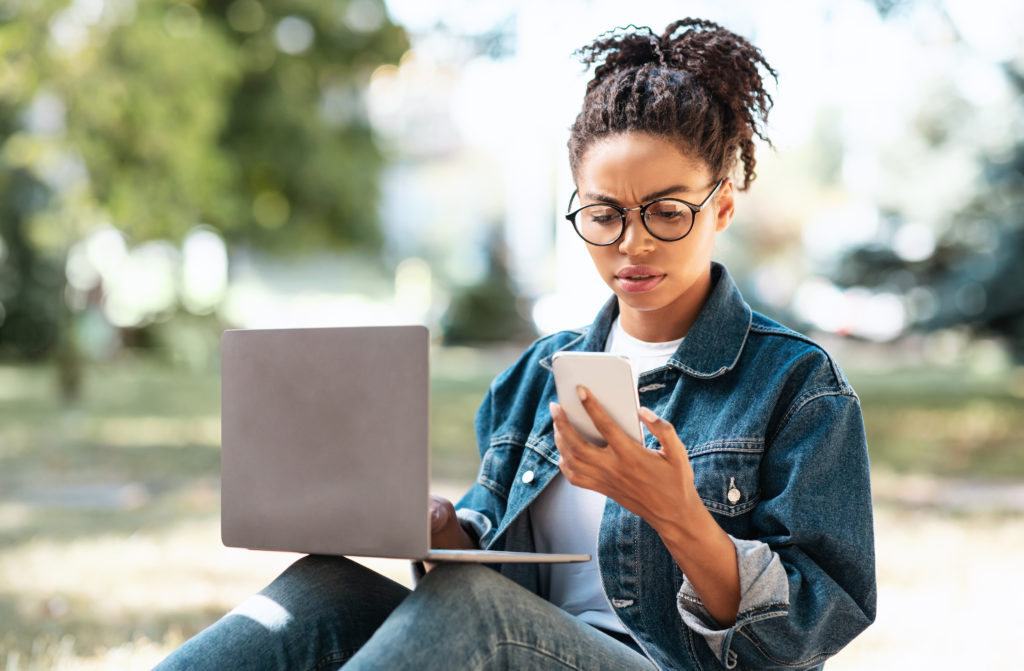 woman sitting outside with laptop while frowning at phone