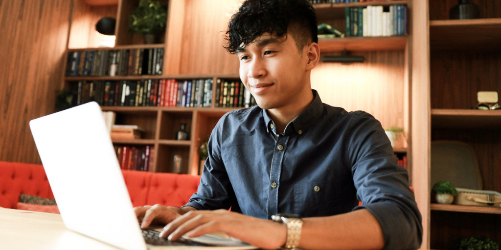 Young man working on tasks using his laptop in a library with books in the background.