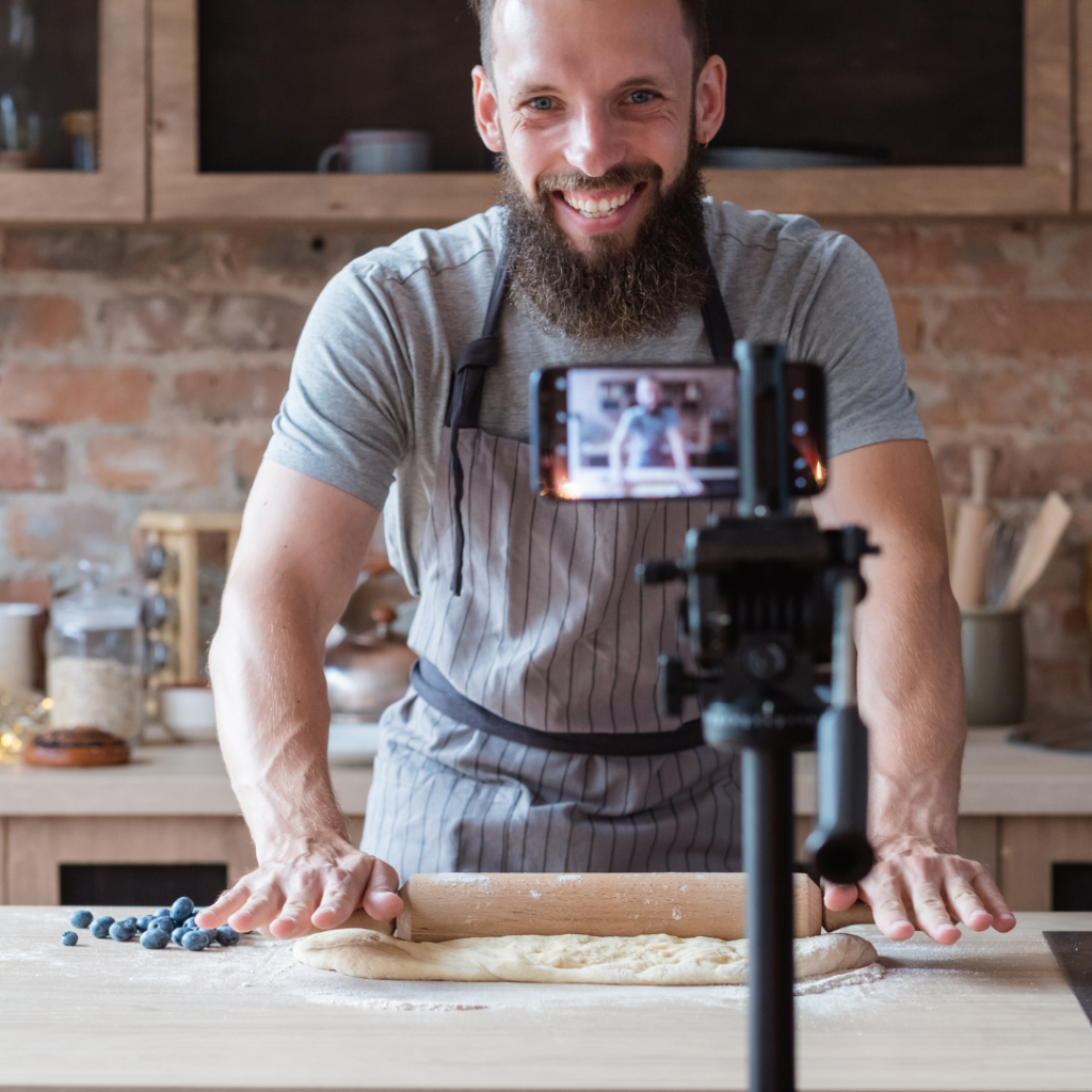 smiling man in kitchen livestreaming as he rolls dough