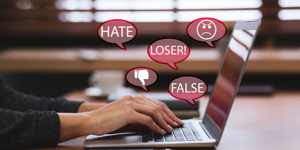  Hands typing on laptop with speech bubbles showing words like hate, loser, and false as well as thumbs down and frowny face. 