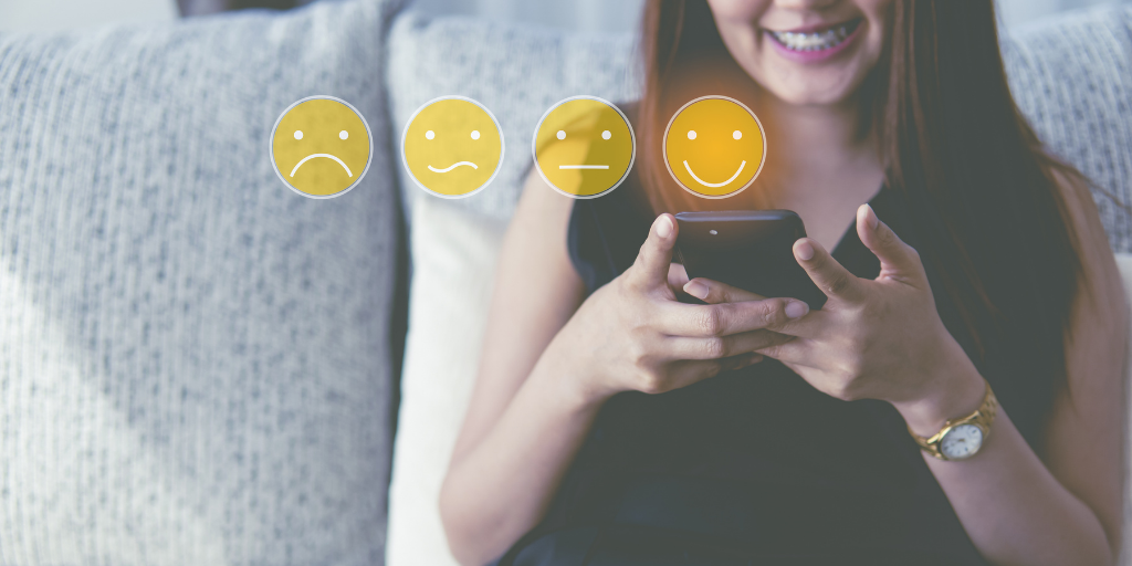 Woman sitting on couch holding smartphone with smile face icon for rating from feedback.