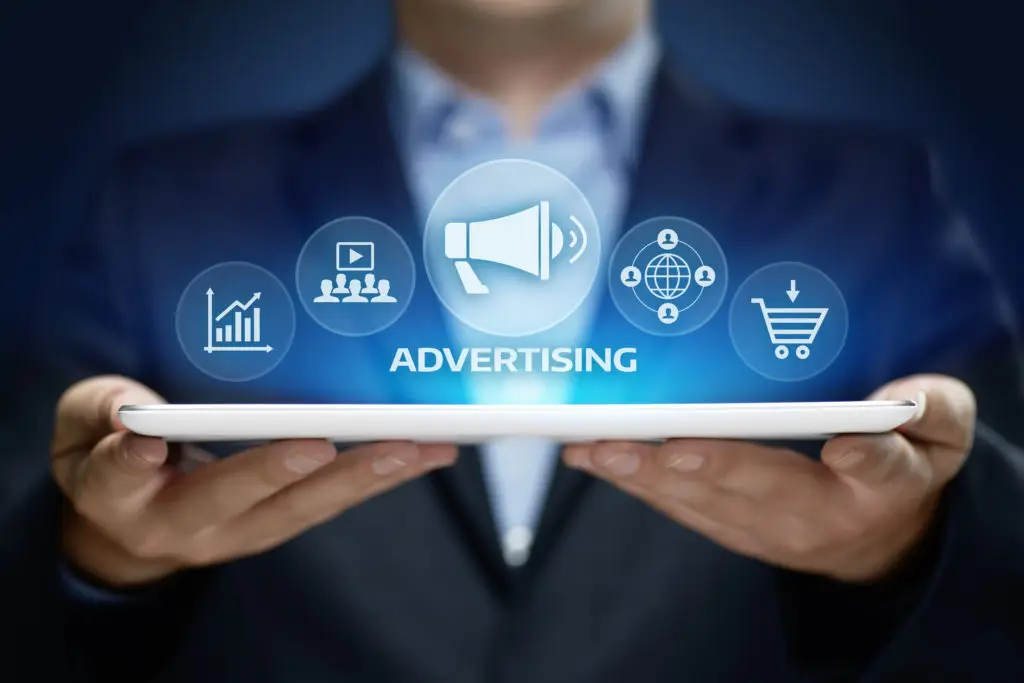 Man holds tablet with stylized graphic symbols above depicting advertising, such as graph, megaphone, and shopping cart.