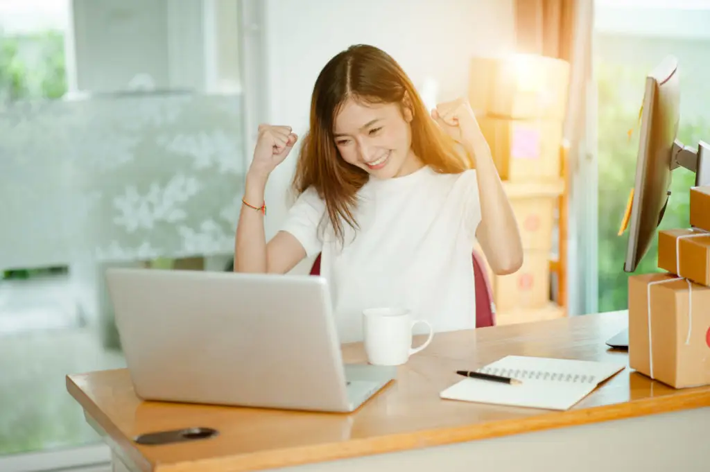 Young woman sitting at laptop and raising hands up to cheer.