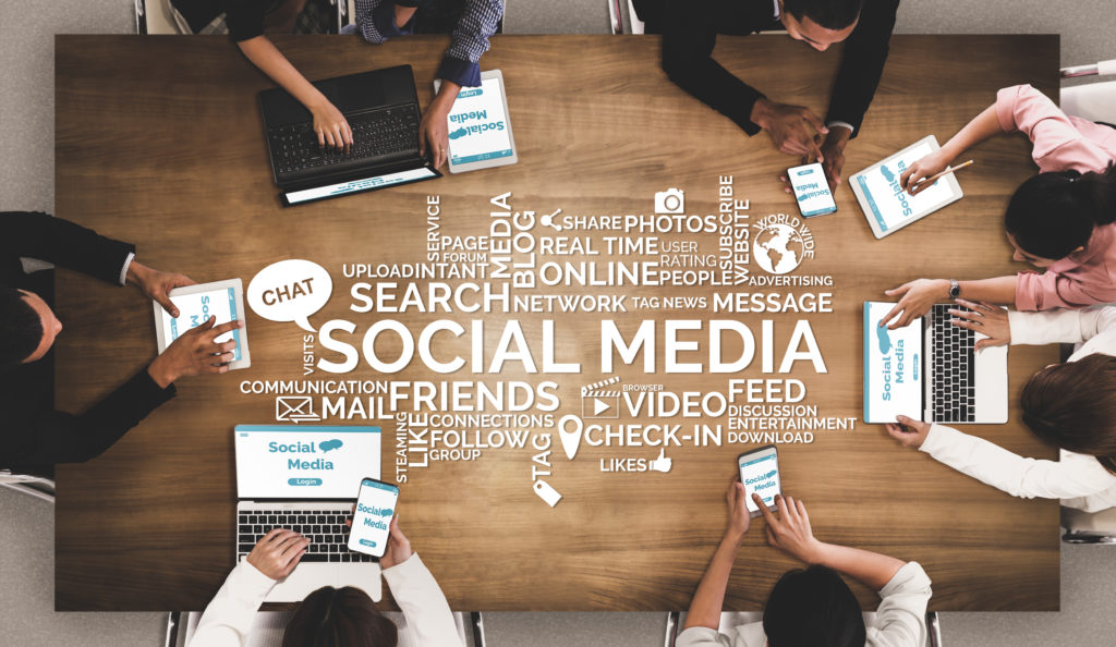  Above view of a group of people sitting at a table working on laptops and mobile devices with social media terms on table.