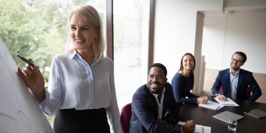 Older businesswoman holding a marker next to a flip board while team members smile on.