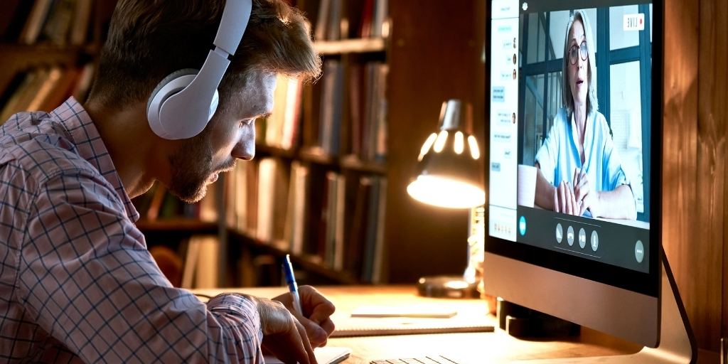 A man wearing headphones takes notes from a remote trainer on his laptop screen during an online class.