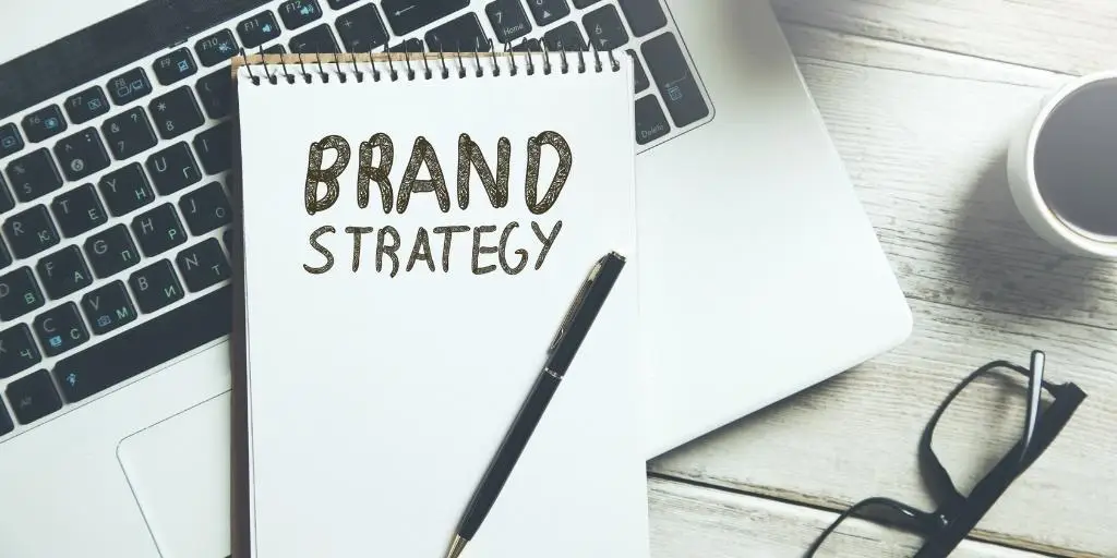 Notepad with the phrase brand strategy resting on laptop keyboard next to glasses.