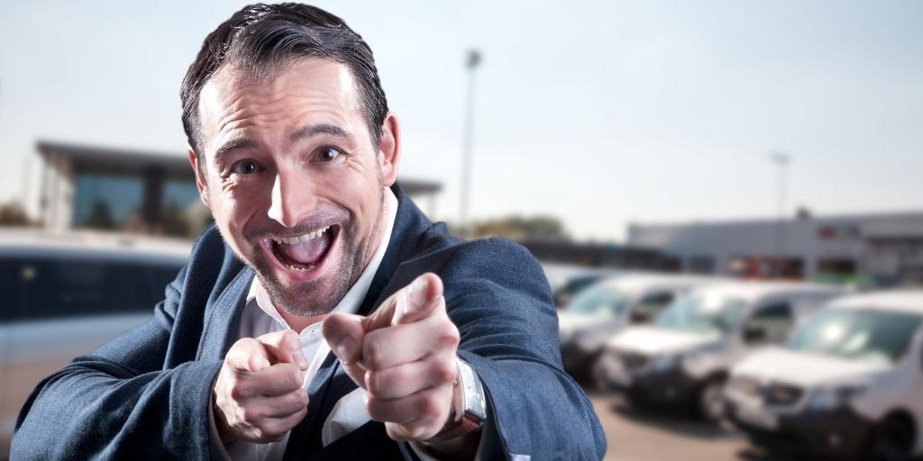 An overenthusiastic car dealer stands in front of a car lot pointing at his customer.