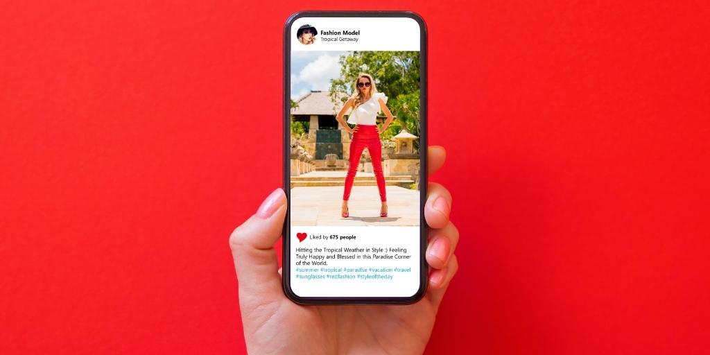 Close-up of hand holding phone displaying an Instagram post of fashion model wearing white top and red pants.