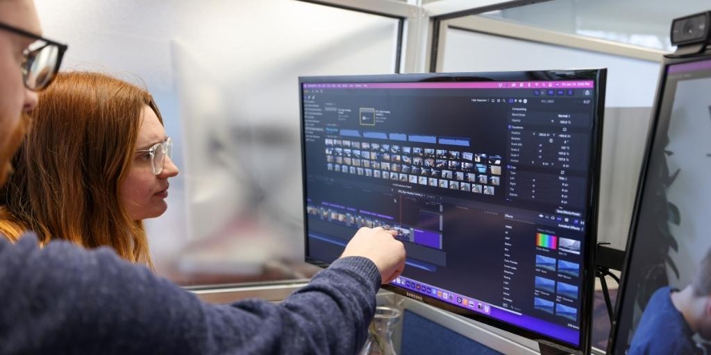 Female editor sitting at computer working on editing video while male colleague points to one of two displays.