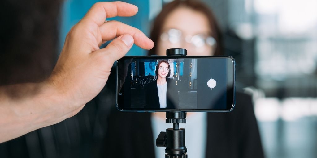 A man’s hand starts a social media video recording of a female professional on a smartphone mounted on a tripod.