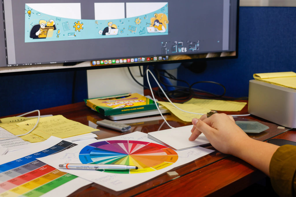 Graphic designer sitting in front of computer displaying a comic in progress and looking at color swatches.