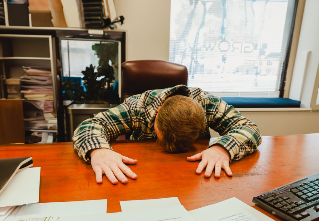 Male employee lying face down on desk, demonstrating overworking employees as a bad management habit.